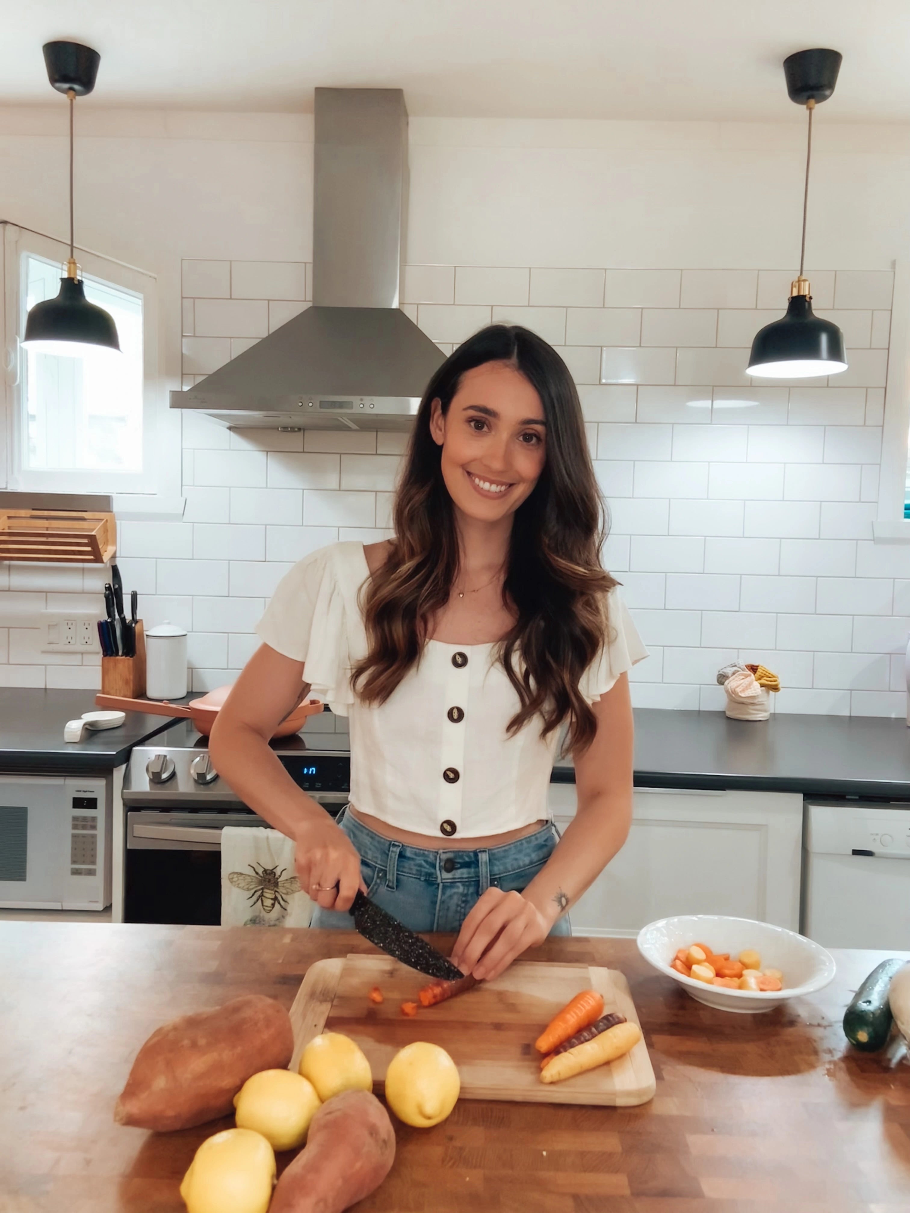 A woman with long hair chops carrots on a wooden board in a modern kitchen. She wears a white top and jeans, and smiles at the camera. The counter has sweet potatoes and lemons, and pendant lights hang from the ceiling.